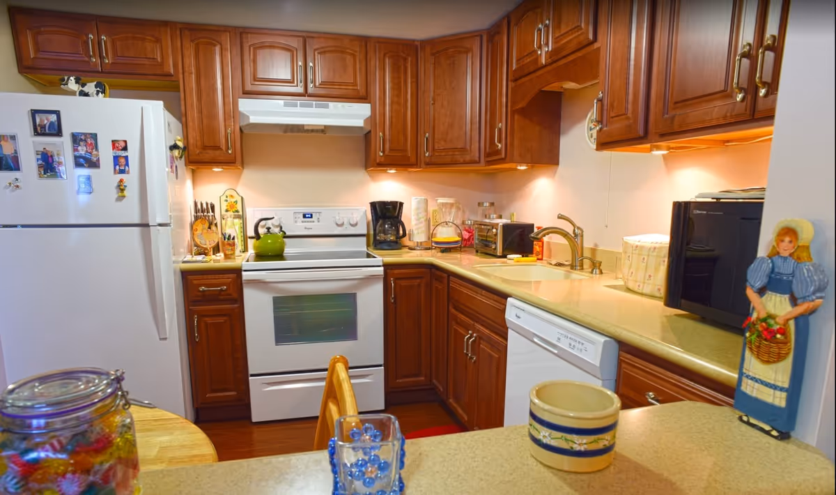 A cozy kitchen with wooden cabinets, a white refrigerator decorated with family photos and magnets, a white stove with a green kettle on top, a coffee maker, a toaster oven, a microwave, and a dishwasher. The countertop has various kitchen items including a paper towel holder and a decorative container. There is a small wooden table with a glass jar of colorful candies and a blue glass cup in the foreground. A decorative wooden figure of a woman holding a basket of flowers is mounted on the wall near the microwave.