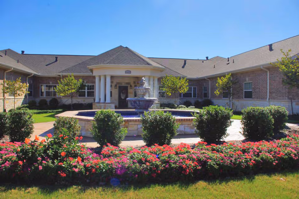 Exterior view of a single-story brick building with a central entrance featuring white columns and a small roof. In front of the entrance is a circular fountain surrounded by neatly trimmed bushes and colorful flower beds under a clear blue sky.