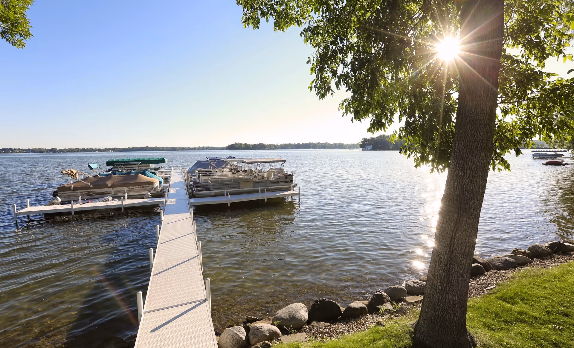 A sunny lakeside scene with a dock extending into the water, several boats moored on either side of the dock, a tree with green leaves on the right side casting shadows, and a rocky shoreline with green grass.