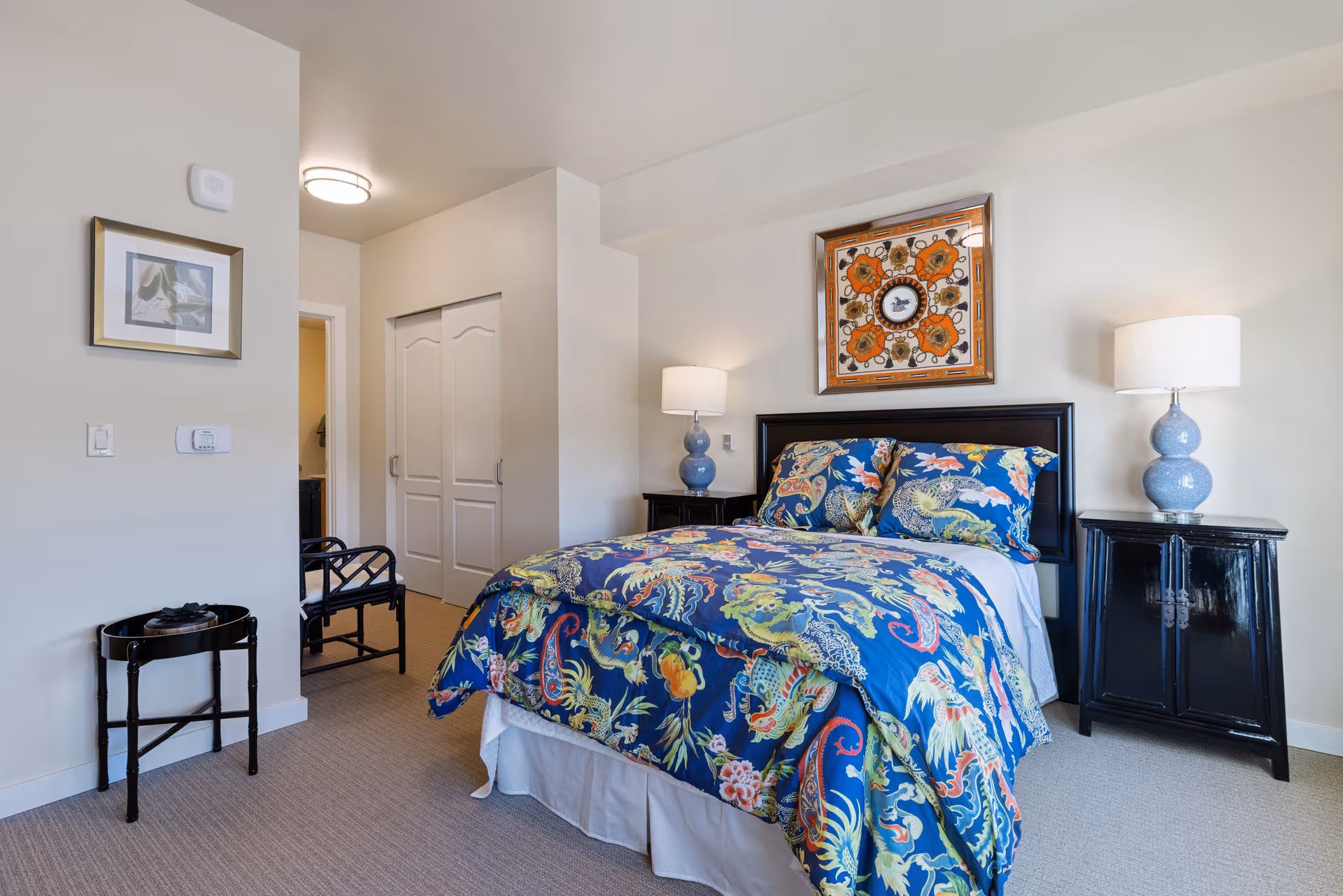 A bedroom with a bed covered in a colorful blue floral and dragon patterned comforter and matching pillows. There are two black nightstands on either side of the bed, each with a blue lamp with a white lampshade. Above the bed hangs a framed artwork with orange and brown tones. The room has beige walls and carpet, a small black table and chair near the entrance, and a closet with white double doors.