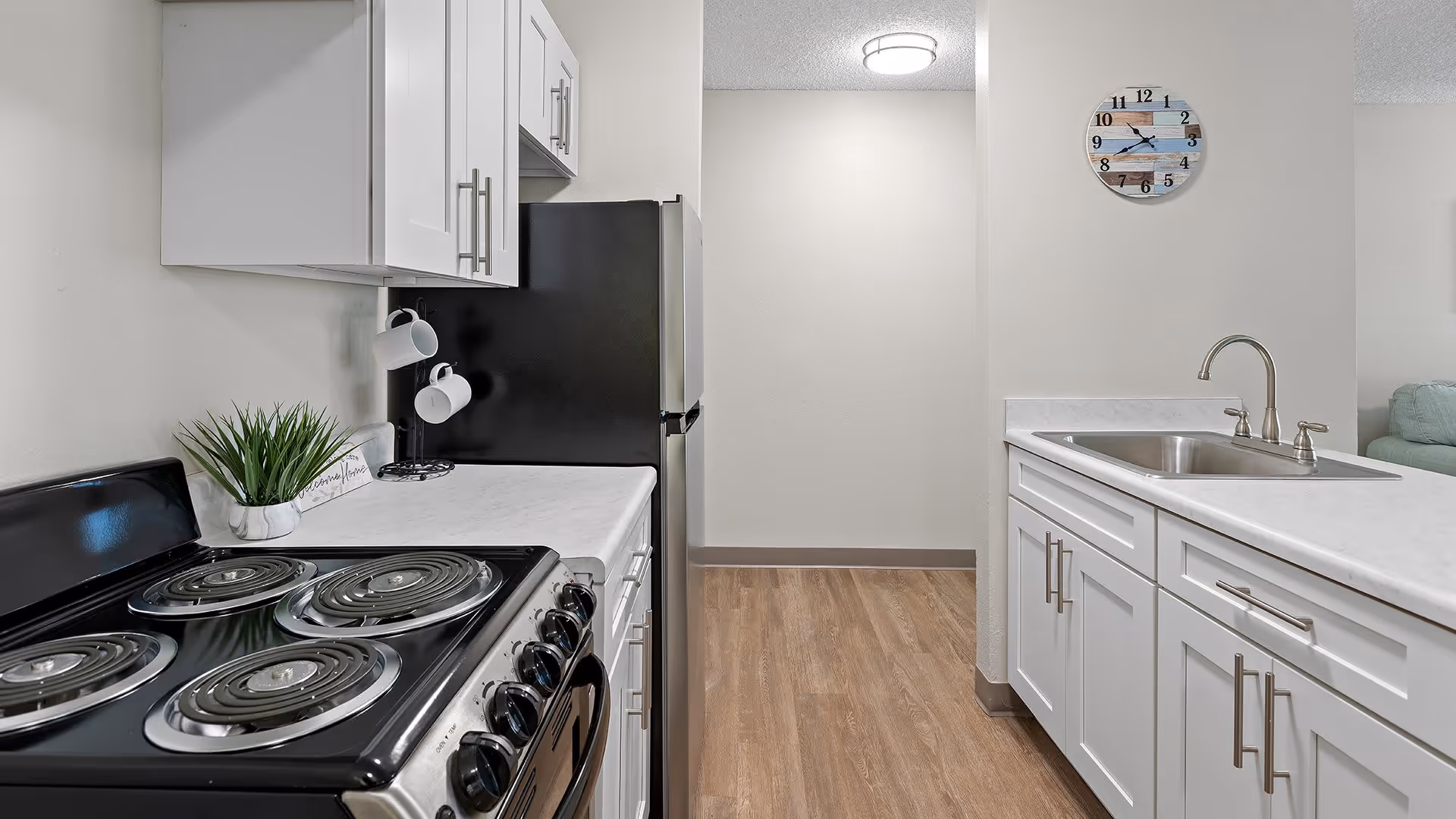 A modern kitchen with white cabinets, a black electric stove with coil burners, a black refrigerator, a white countertop with a sink, a small green plant, two white mugs hanging on hooks, and a wall clock with a beach-themed design. The floor is wood, and the walls are light-colored.