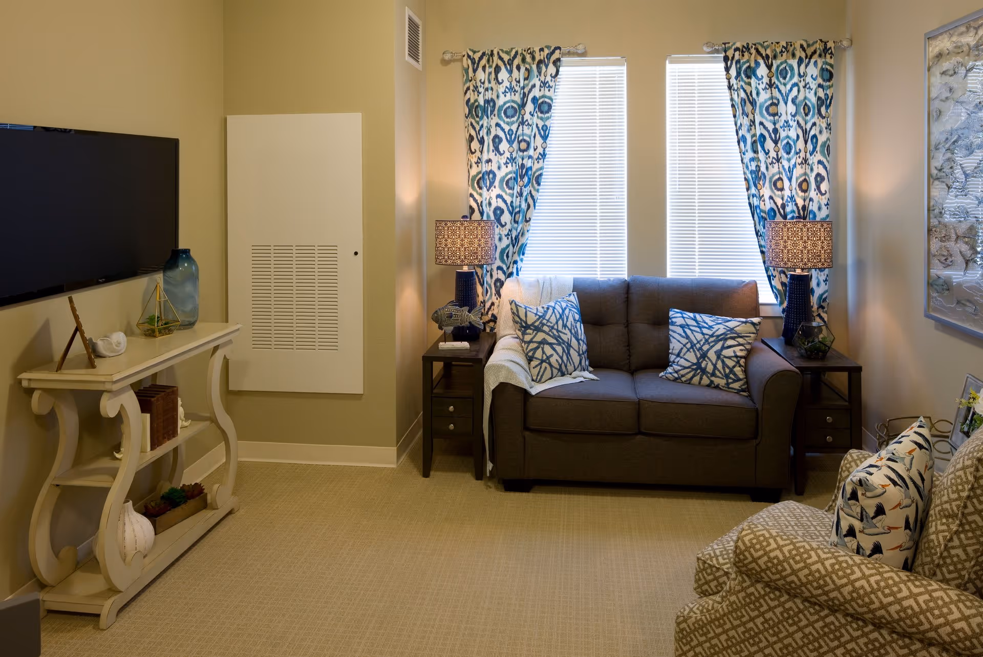 A cozy living room with a gray loveseat adorned with two blue and white patterned pillows, flanked by two dark wooden side tables each with a decorative lamp. Two windows behind the loveseat have white blinds and blue patterned curtains. To the right, there is a patterned armchair with a pillow featuring bird designs. On the left wall, a flat-screen TV is mounted above a white console table decorated with a blue vase, a small sculpture, and other decorative items. The room has beige walls and carpeted flooring.