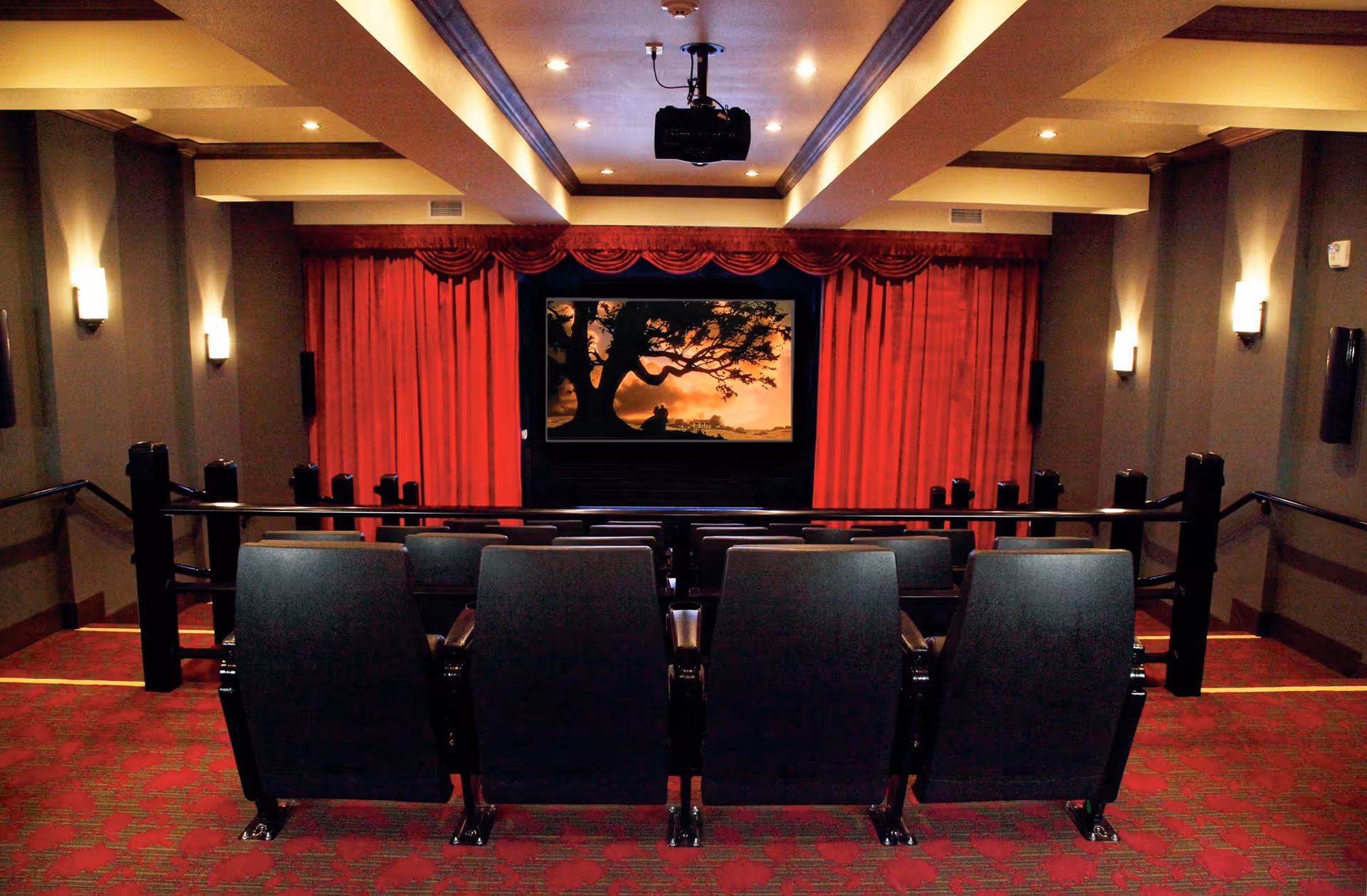 Interior view of a small theater room with red carpet and red curtains framing a large screen displaying an image of a tree silhouette at sunset. There are two rows of black theater seats facing the screen, and the room is softly lit with wall sconces and ceiling lights.
