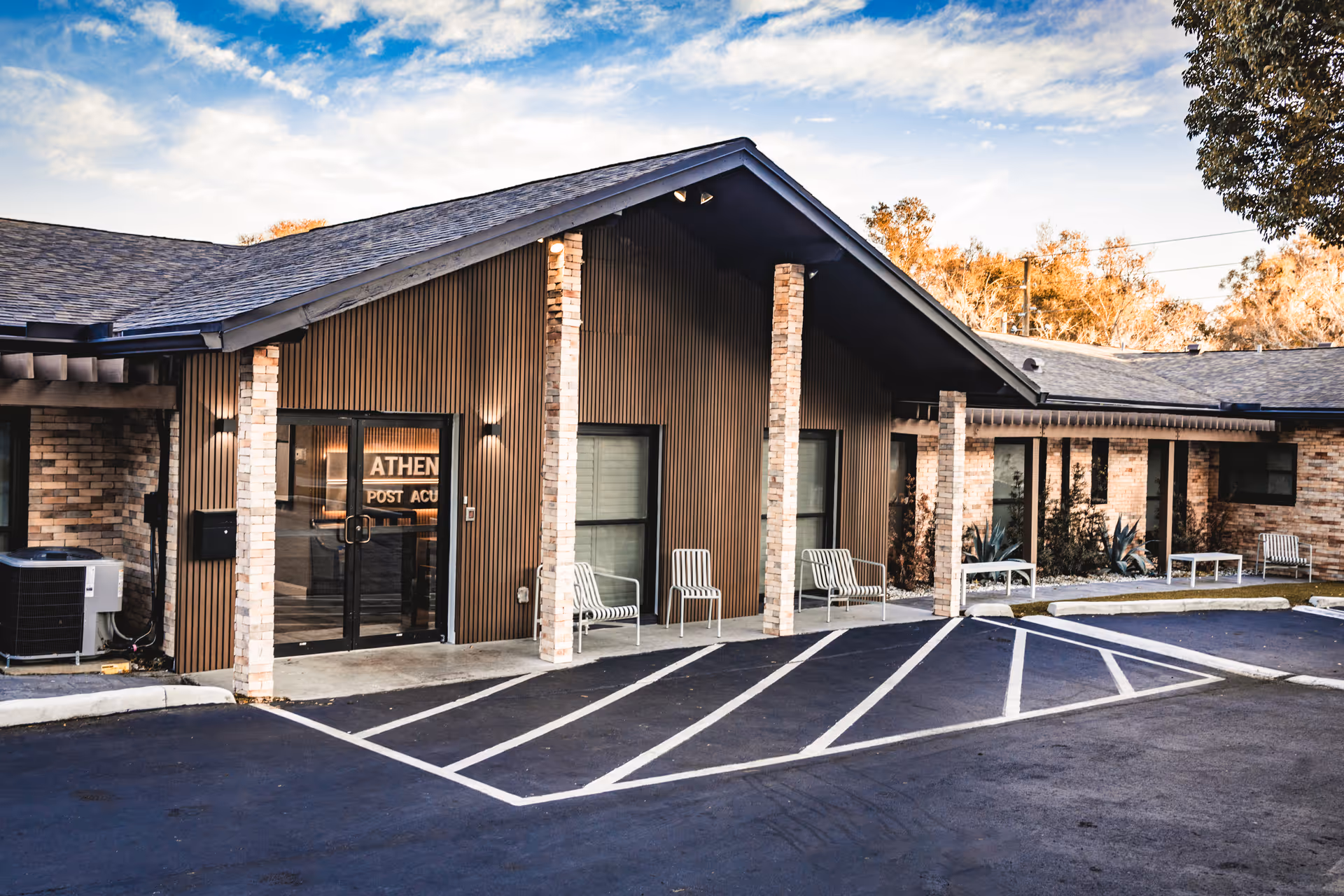 Front exterior of the Athens Post Acute single-story building with a covered entrance, brick columns, chairs, and marked parking spaces.