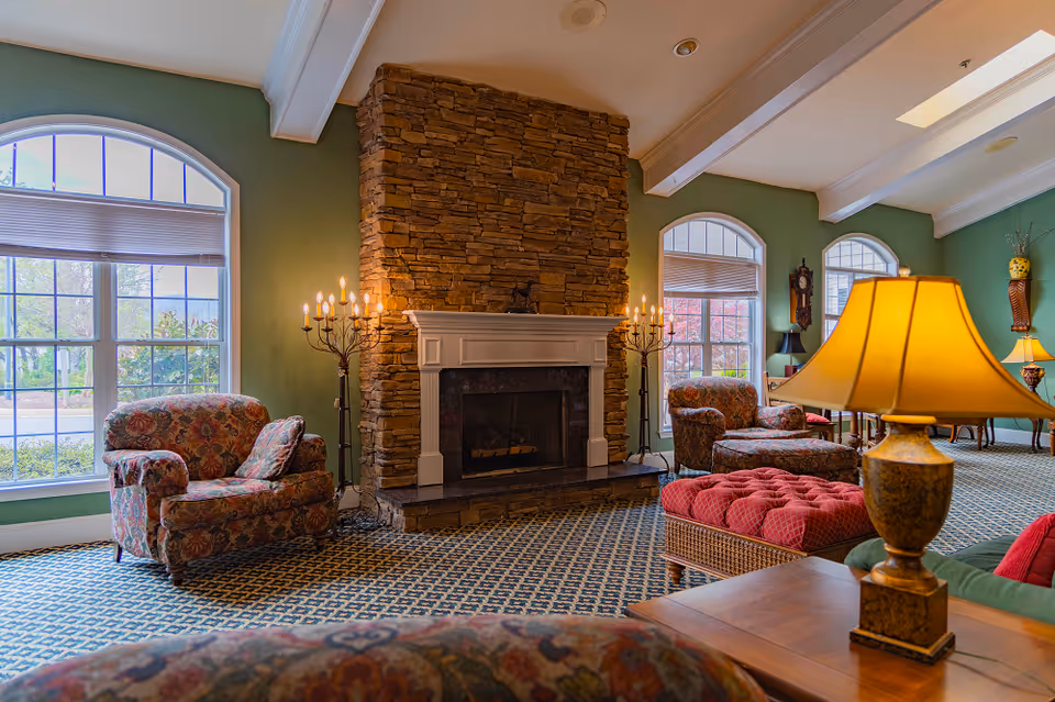 A cozy senior living room with green walls, large arched windows, and a stone fireplace in the center. The room is furnished with floral upholstered armchairs, a red tufted ottoman, and a wooden table with a lamp. Two candelabra-style floor lamps flank the fireplace, and the carpet has a blue and white pattern.