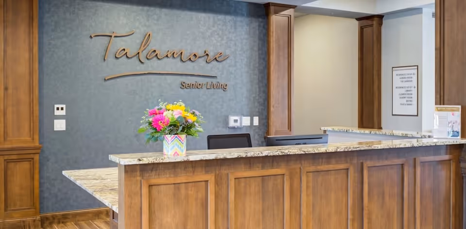 Lobby reception desk with a granite countertop, wooden paneling, a colorful flower arrangement, and 'Talamore Senior Living' wall signage.