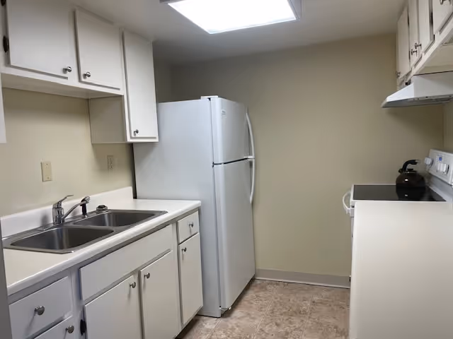 A small kitchen with white cabinets, a double sink, a white refrigerator, a white stove with a kettle on it, and beige tiled flooring. The walls are painted light beige and there is a fluorescent ceiling light.