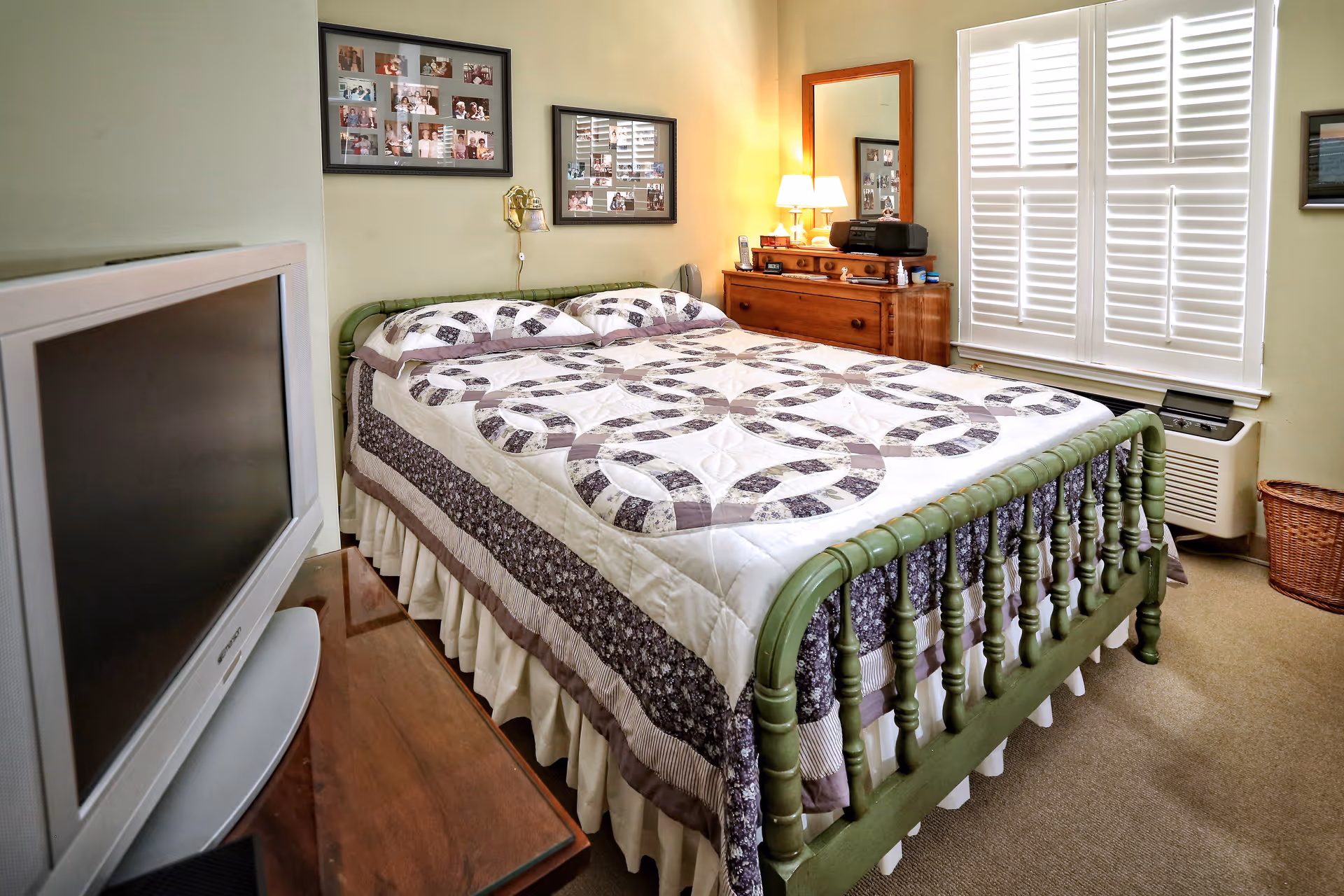 A neatly made bedroom with a patterned quilt on a green wooden bedframe, dresser with lamp and mirror, TV, and shuttered window.