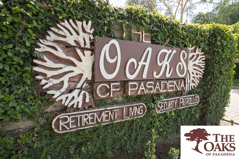 A large outdoor sign for Oaks of Pasadena retirement living, mounted on a wall covered with green ivy. The sign features the text 'OAKS OF PASADENA RETIREMENT LIVING SECURITY PATROLLED' with decorative leaf designs on either side. There is also a smaller logo in the bottom right corner showing a tree and the text 'THE OAKS OF PASADENA'.
