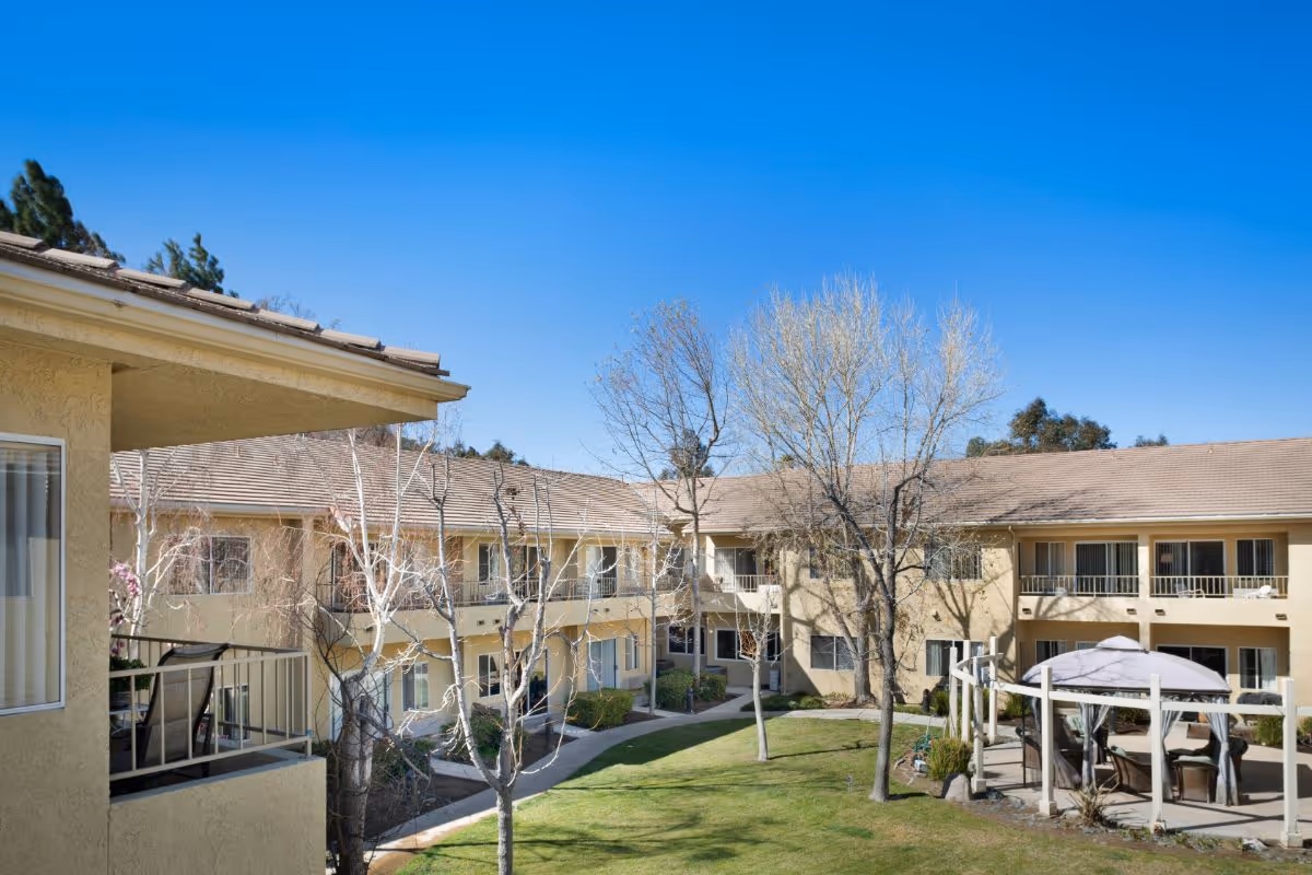 Outdoor courtyard area of a senior living facility with a two-story beige building surrounding a grassy lawn. There are leafless trees, a paved walkway, and a covered seating area with chairs and a table under a gazebo. The sky is clear and blue.