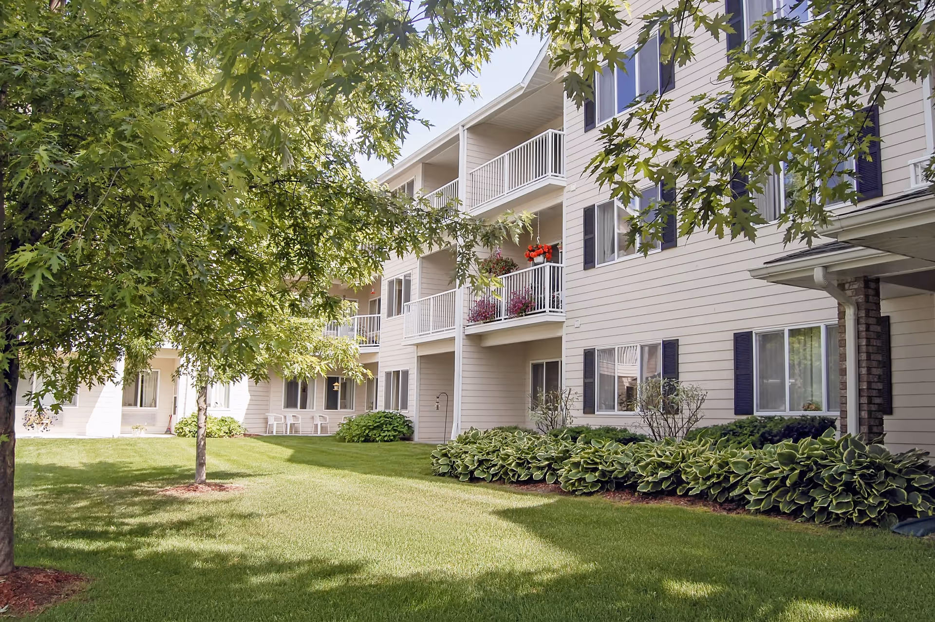 Exterior view of a multi-story senior living facility building with balconies, surrounded by green grass, trees, and shrubs under a clear sky.