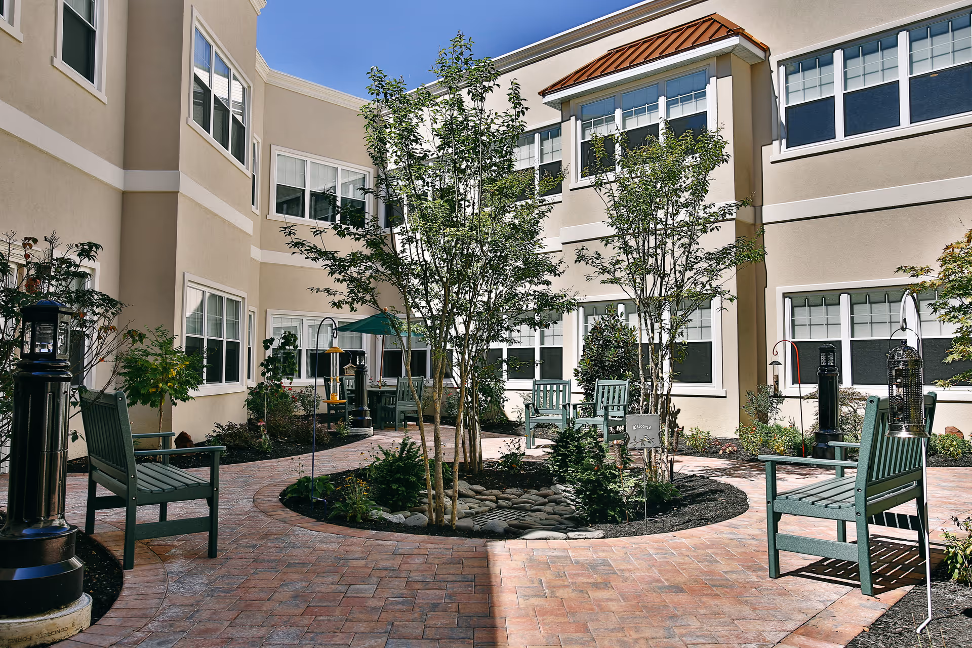 Sunny landscaped courtyard with brick pathways, benches, small trees, and the surrounding two-story building.