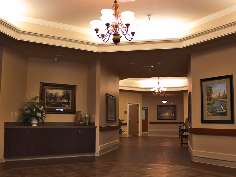 Interior hallway of a senior living facility with beige walls, dark wood flooring, framed paintings on the walls, a decorative chandelier on the ceiling, and a cabinet with a flower arrangement and vases.