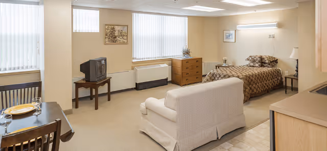 A cozy studio apartment with a single bed covered in a brown patterned bedspread, a beige loveseat facing a small TV on a wooden stand, a wooden dresser, and a dining table with two chairs set with plates and glasses. The room has large windows with vertical blinds and neutral-colored walls and carpet.