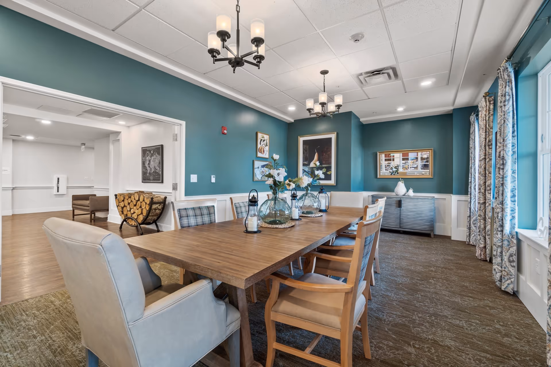 A well-lit dining room with a long wooden table surrounded by various chairs, including cushioned armchairs and wooden chairs with plaid cushions. The walls are painted teal with white wainscoting, decorated with framed artwork. Two chandeliers hang from the ceiling, and large windows with patterned curtains allow natural light to enter. In the background, there is an open doorway leading to another room with wooden flooring and a stack of firewood in a holder.