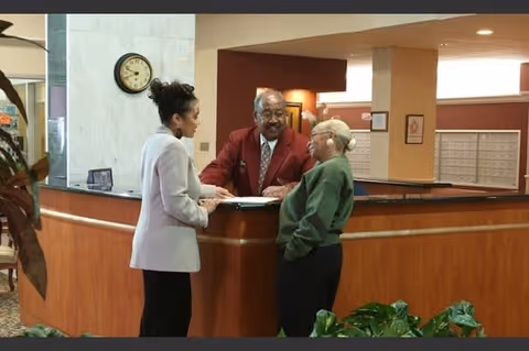 Three people standing and talking at a reception desk in an indoor setting. One person is behind the desk wearing a red jacket, while two others stand in front of the desk engaged in conversation. There is a clock on the wall and some plants in the foreground.