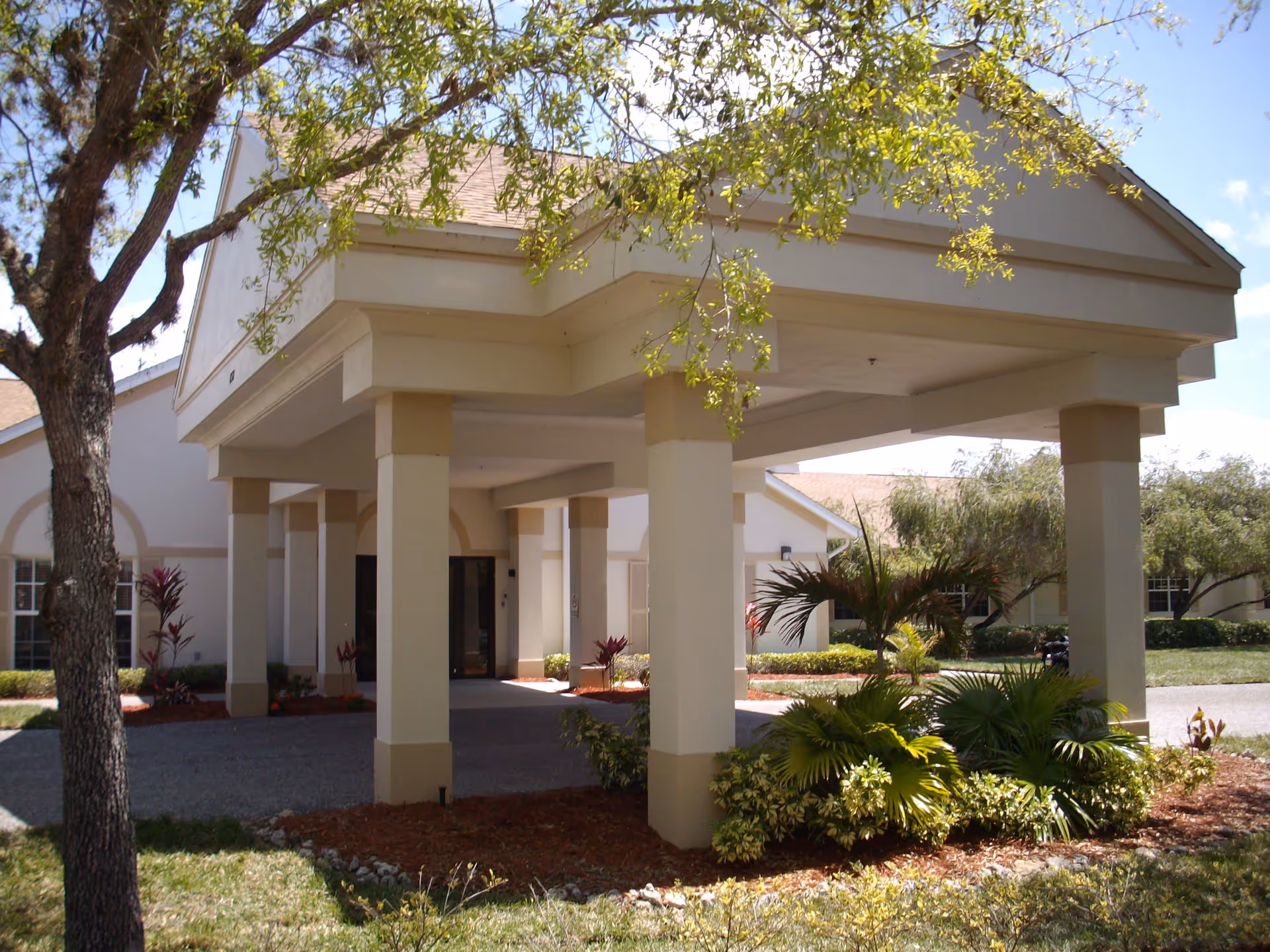 Exterior view of the entrance to Sunset Lake Health and Rehabilitation Center showing a covered driveway supported by columns, surrounded by landscaped greenery and trees under a clear sky.