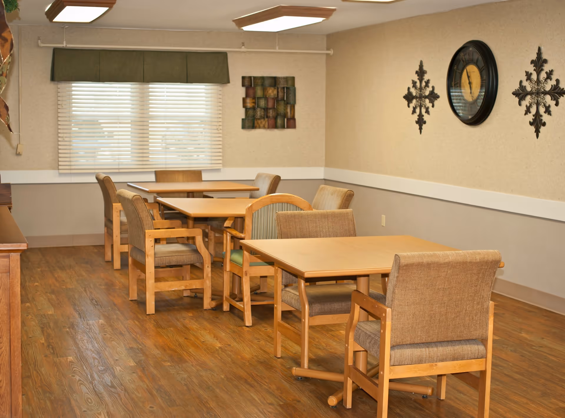 A dining room with wooden tables and cushioned chairs arranged neatly on a wooden floor. The room has beige walls with decorative wall art and a clock. A window with blinds and a green valance lets in natural light.