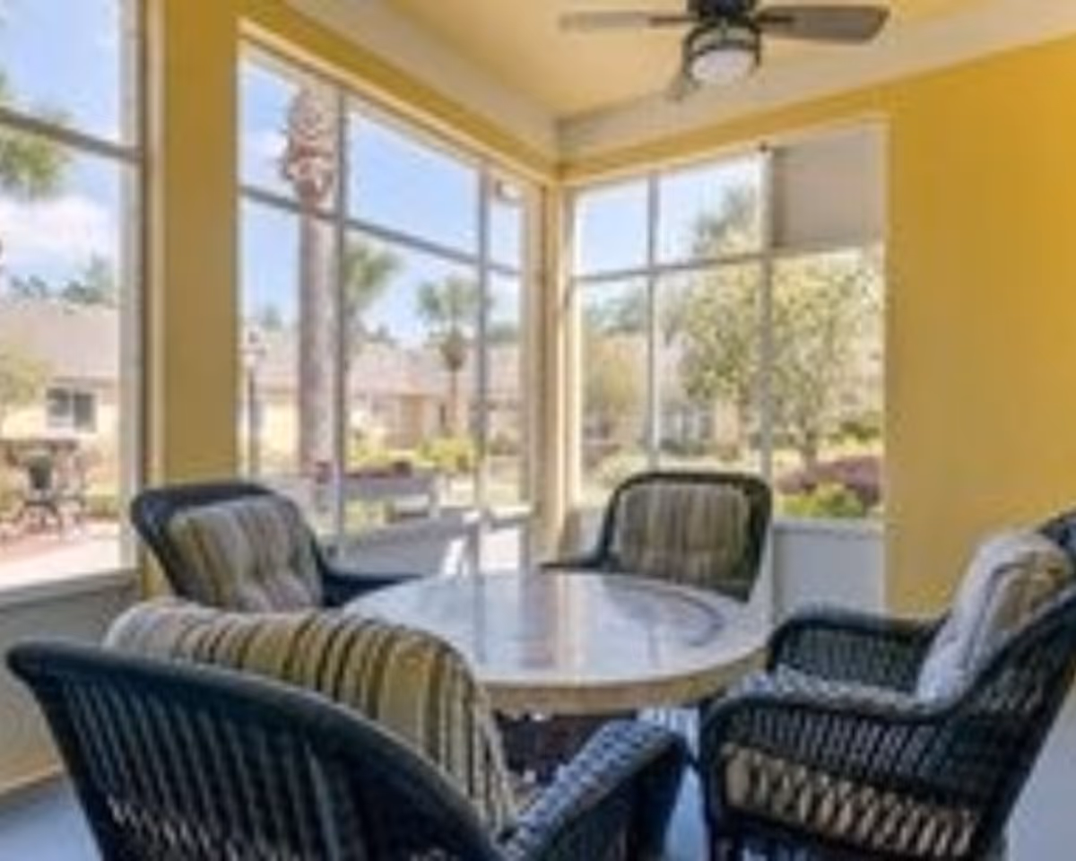 A bright sunroom with large windows showing an outdoor view of trees and houses. The room has yellow walls and contains a round table surrounded by four cushioned wicker chairs. A ceiling fan with a light fixture is mounted on the ceiling.