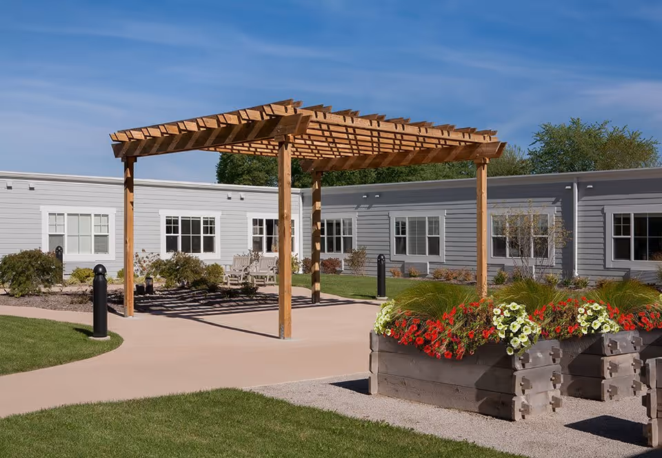 Outdoor courtyard area at StoryPoint Schererville featuring a wooden pergola over a paved walkway, surrounded by green grass, flower beds with red and white flowers, and a gray building with multiple windows in the background under a clear blue sky.
