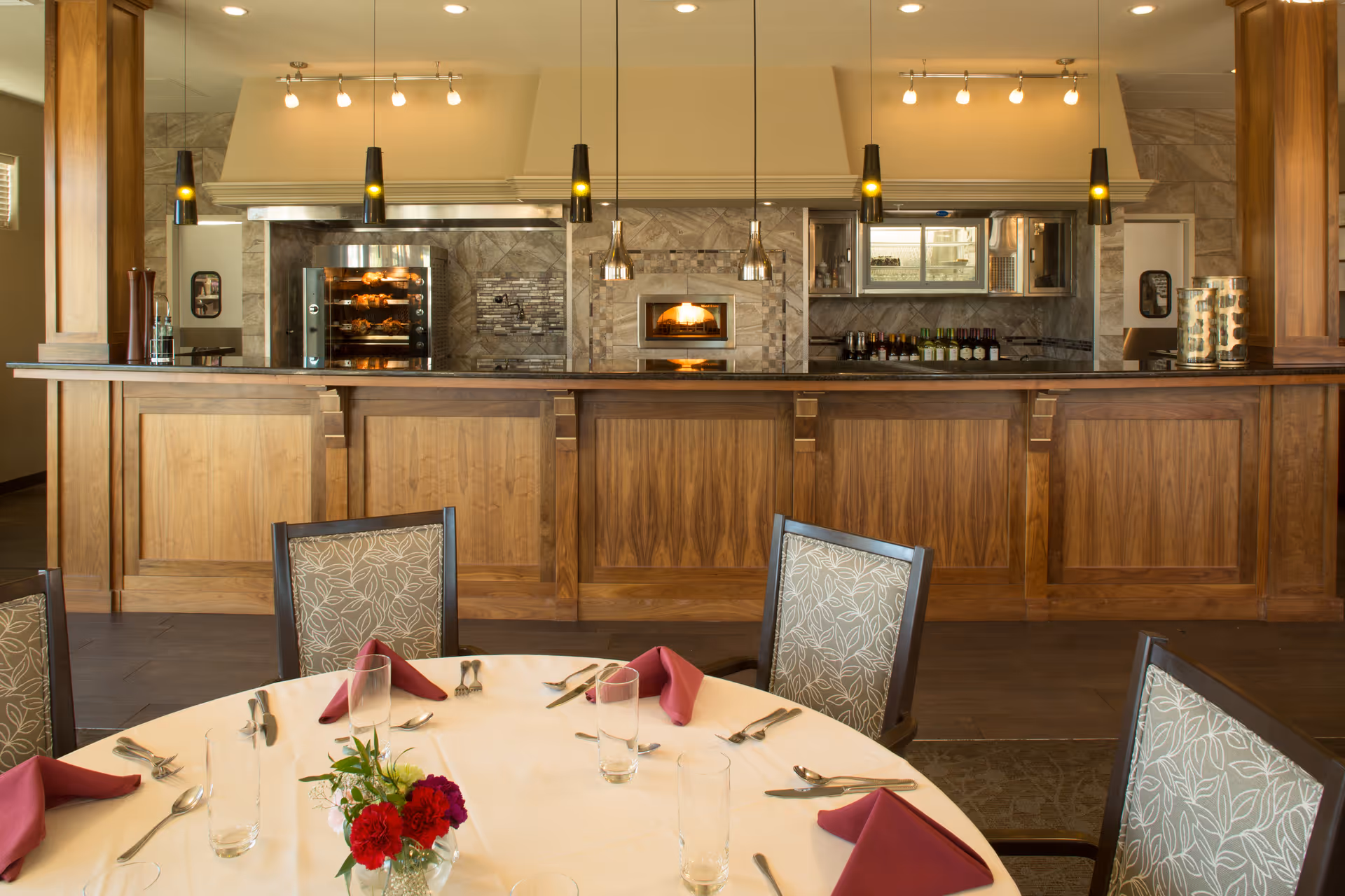 A round dining table set with napkins and a small floral centerpiece in front of a wooden service counter and kitchen area with pendant lights.