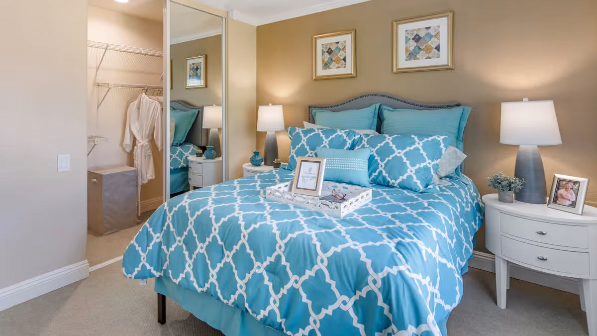 A neatly made bed with a blue and white patterned comforter and multiple pillows in a bedroom. There are two bedside tables with lamps, a small plant, and framed photos. A tray with a framed picture and glasses is placed on the bed. The room has beige walls, two framed artworks above the bed, and a closet with a white robe hanging inside, visible through a mirrored sliding door.