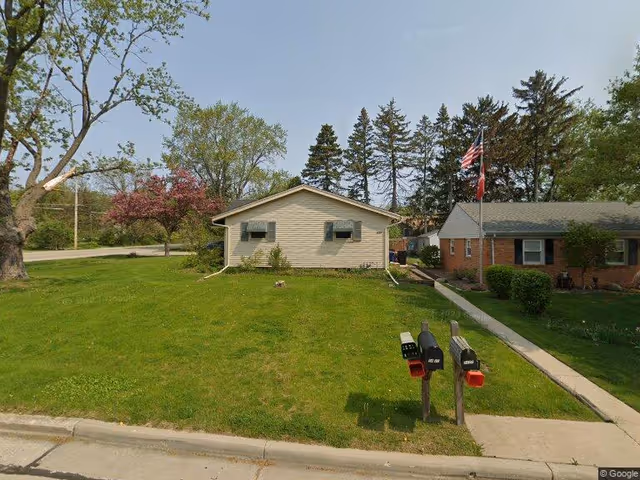 Single-story residential building with a grassy front yard, two mailboxes in the foreground and a neighboring brick house with a flag.