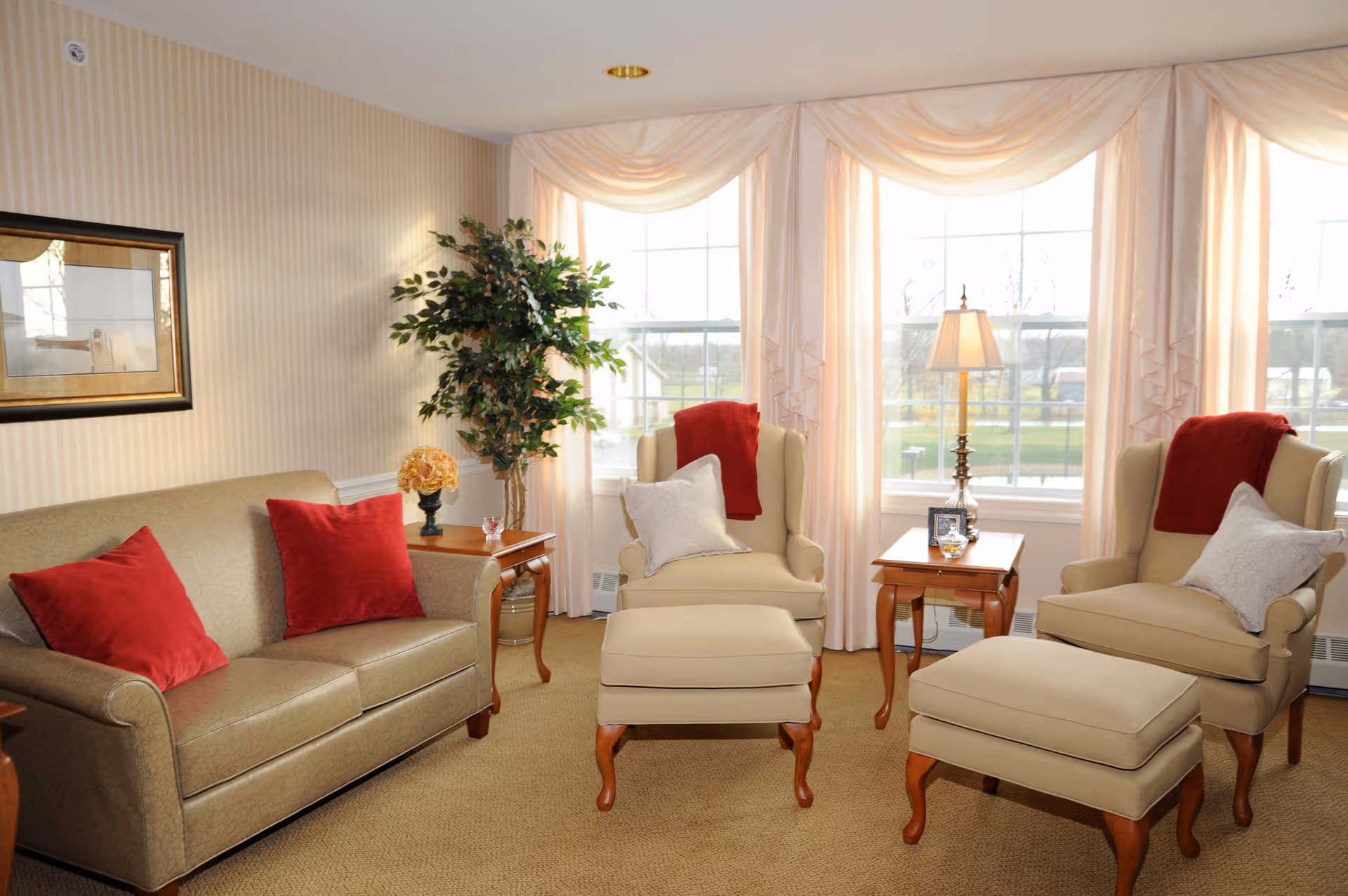 Bright living room with beige sofa and matching armchairs and ottomans, red accent pillows, side tables, a lamp, and large draped windows.