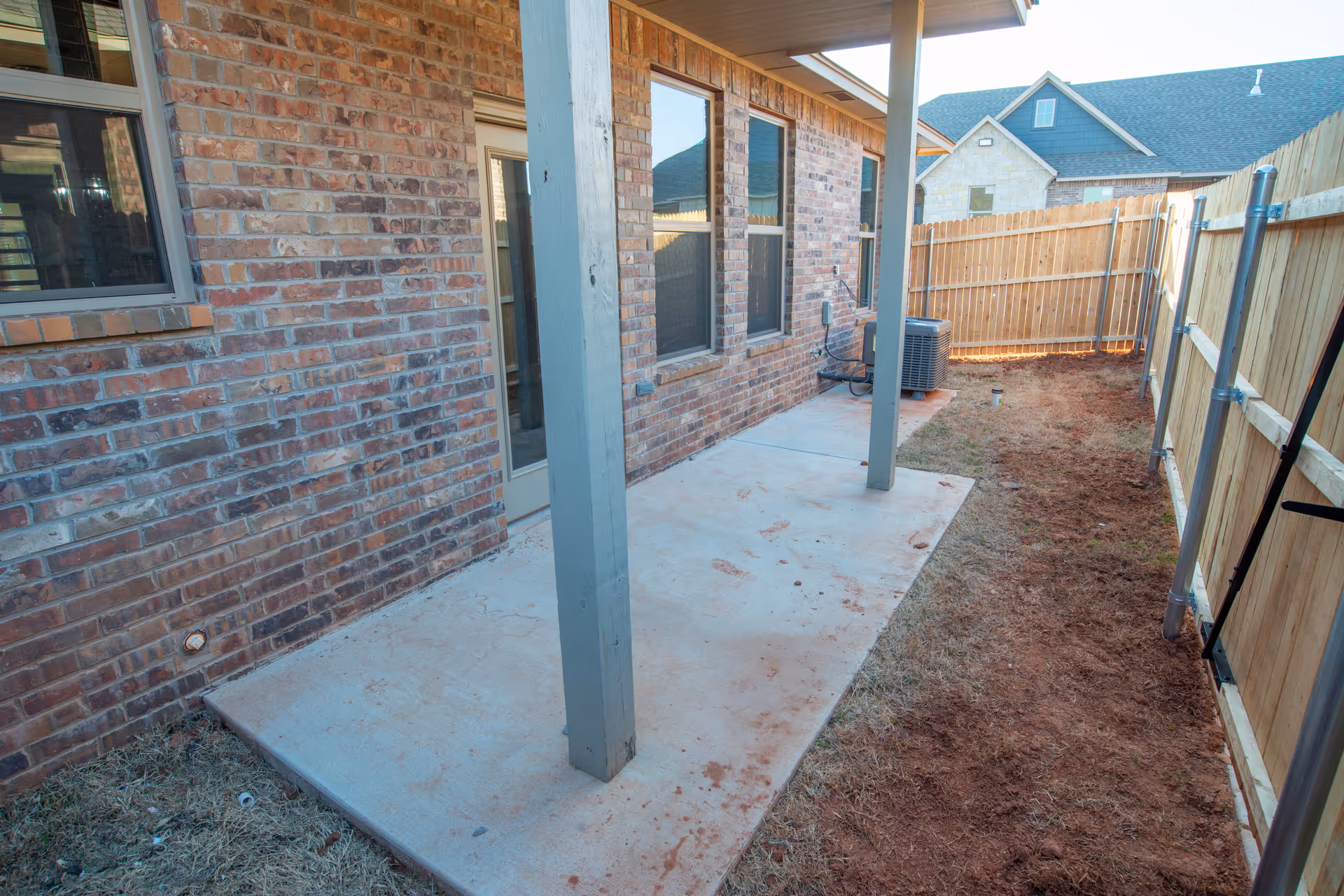 A narrow outdoor patio area with a concrete floor adjacent to a brick wall of a building. The patio is supported by two vertical wooden posts and is bordered by a wooden fence on the right side. There is an air conditioning unit near the far end of the patio and neighboring houses are visible in the background.