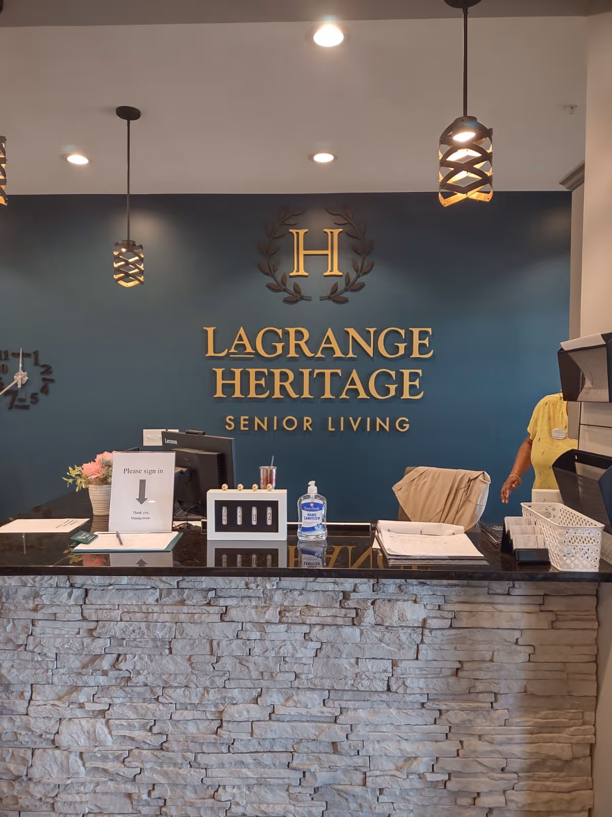 Reception desk area of LaGrange Heritage Senior Living with a stone facade counter, a dark blue wall behind displaying the facility's name and logo in gold letters, two pendant lights hanging from the ceiling, a clock on the wall, a computer monitor, hand sanitizer, and a sign requesting visitors to sign in.