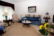 A cozy living room with a blue tufted sofa, two floral patterned armchairs, a round wooden coffee table with decorative items, a side table with a lamp, and a window with sheer white curtains and dark valance. There is a framed picture on the wall and a vase with yellow flowers in the foreground.