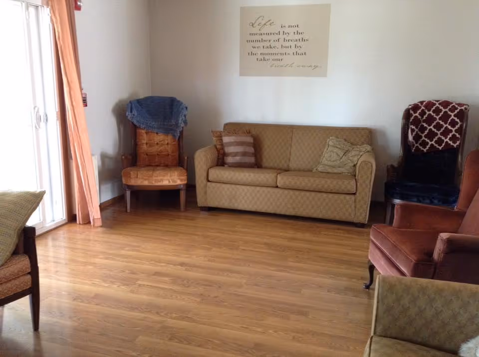A living room with wooden flooring featuring a beige sofa with two cushions, an orange armchair, a brown cushioned chair with a blue throw draped over it, and a dark chair with a red and white patterned blanket. There is a sliding glass door with peach-colored curtains on the left side and a wall decoration with an inspirational quote above the sofa.