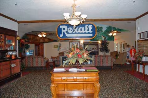 Interior view of a senior living facility lobby or common area with a wooden table holding a floral arrangement in the center. A large hanging sign reads 'Rexall'. The room has carpeted floors, ceiling fans, and various seating areas with chairs and tables. Two people are visible on the right side near a wall with framed pictures.