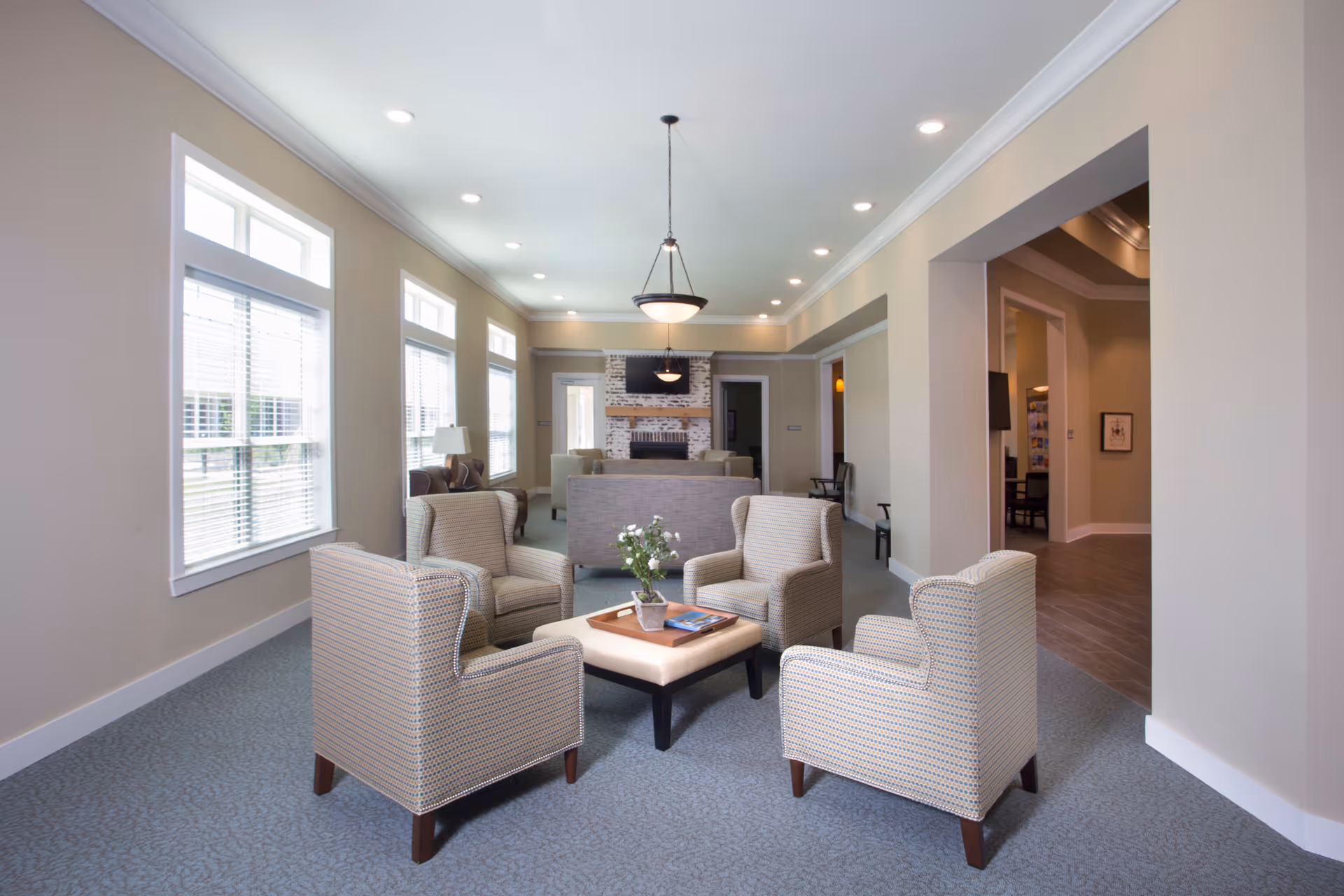A bright and spacious living room area with four patterned armchairs arranged around a square ottoman with a tray and a small plant on it. The room has large windows with blinds on the left side, recessed ceiling lights, a hanging light fixture, and a white brick fireplace with a mounted TV above it in the background. The walls are painted beige and the floor is carpeted in a light blue pattern.