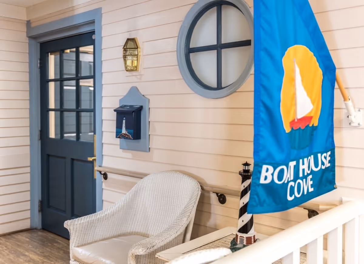 Indoor seating area with a white wicker chair, a blue door with glass panes, a round window with blue trim, a wall-mounted mailbox with a lighthouse image, a brass wall light fixture, and a blue flag with a sailboat and the words 'BOAT HOUSE COVE'.