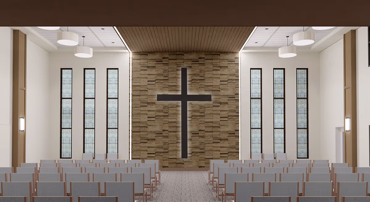 Interior view of a chapel or worship room with rows of chairs facing a stone wall featuring a large illuminated cross. The room has tall vertical windows with frosted glass on both sides and modern ceiling lights.