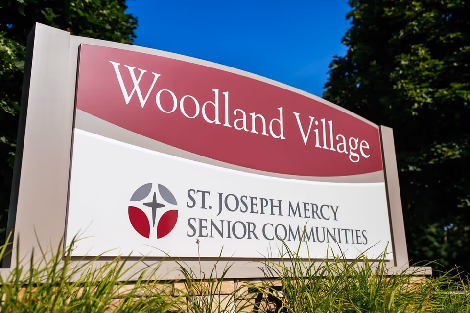 Outdoor sign for Woodland Village, part of St. Joseph Mercy Senior Communities, with a clear blue sky and greenery in the background.