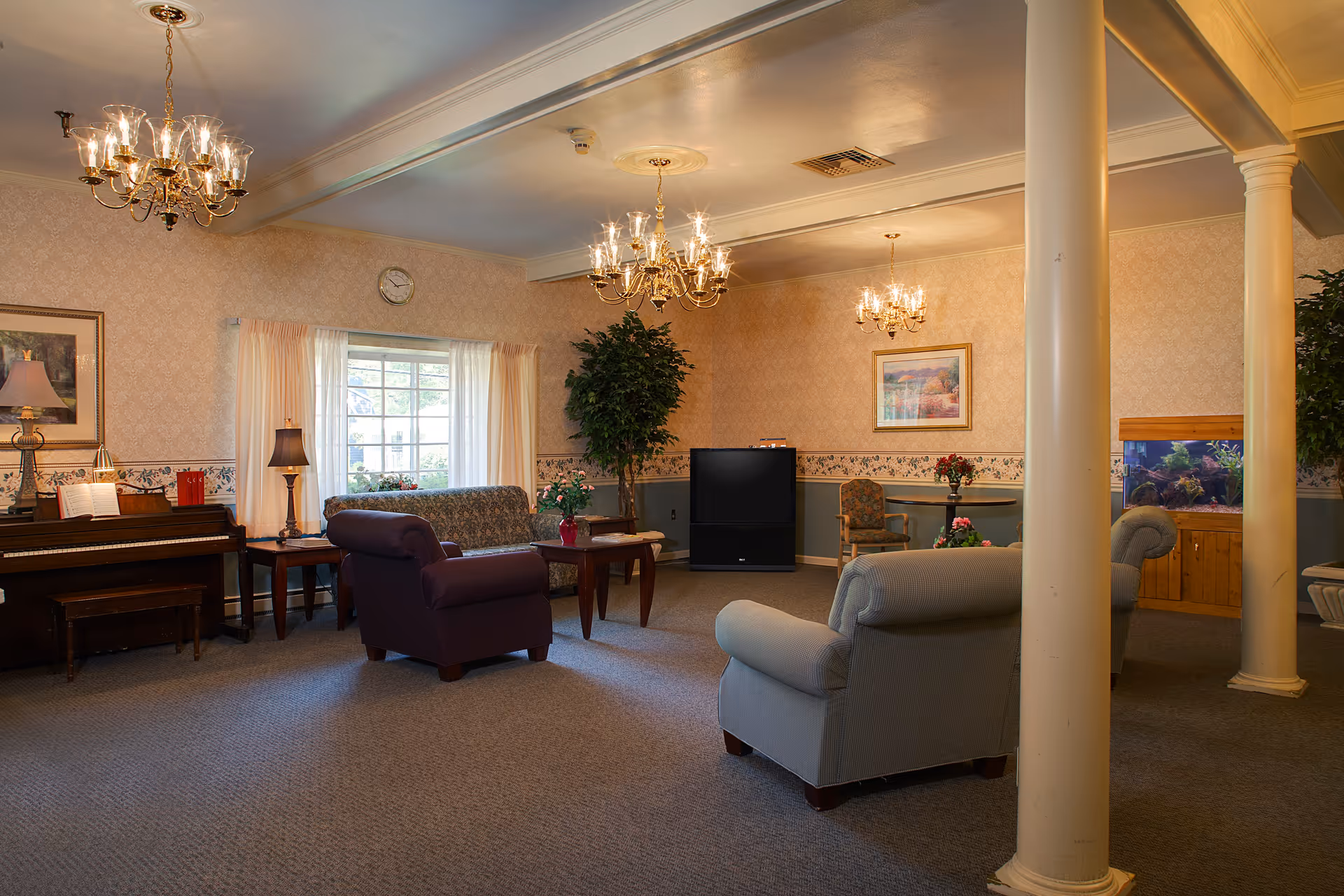 Comfortable senior living common room with armchairs, a sofa, piano, TV, and an aquarium under chandeliers.