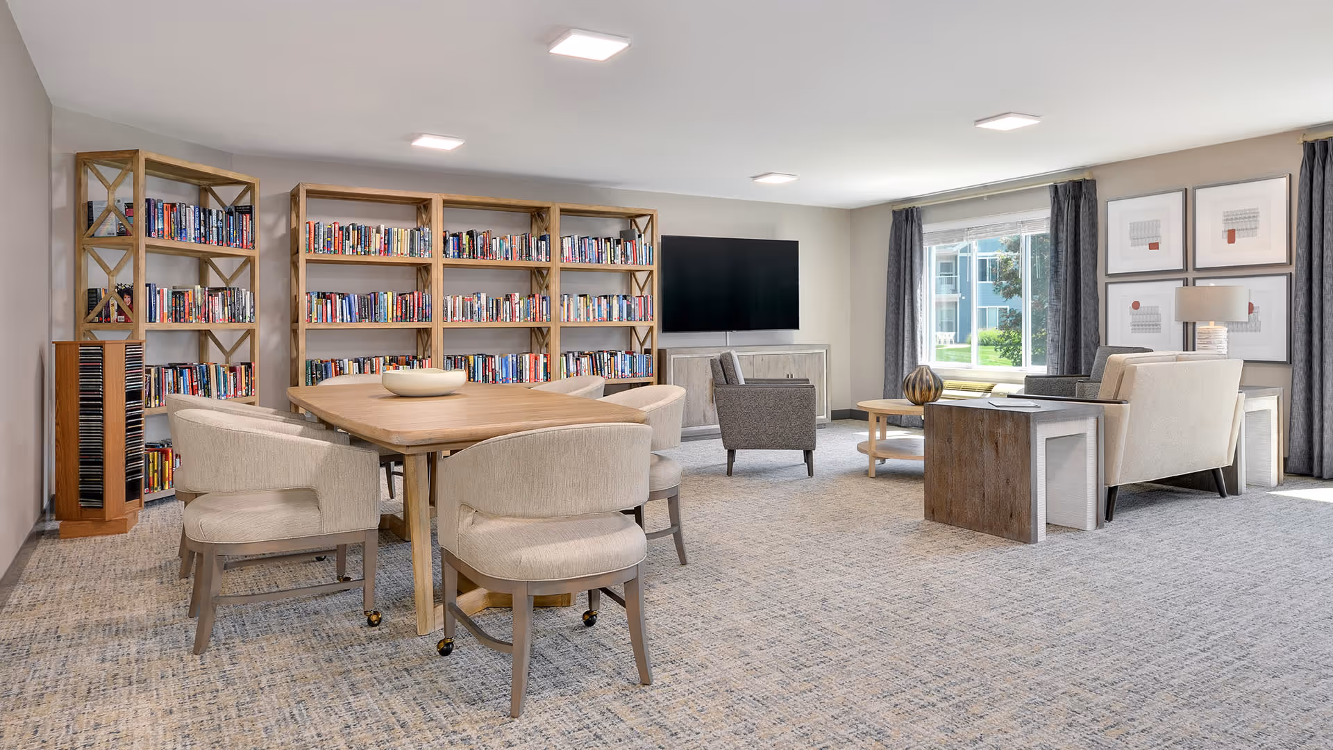 A bright and spacious common area in a senior living facility featuring a wooden table with six beige upholstered chairs on casters, a large bookshelf filled with books, a flat-screen TV mounted on the wall, and a seating area with armchairs and a sofa near a window with gray curtains. The room has light-colored walls and carpeted flooring.