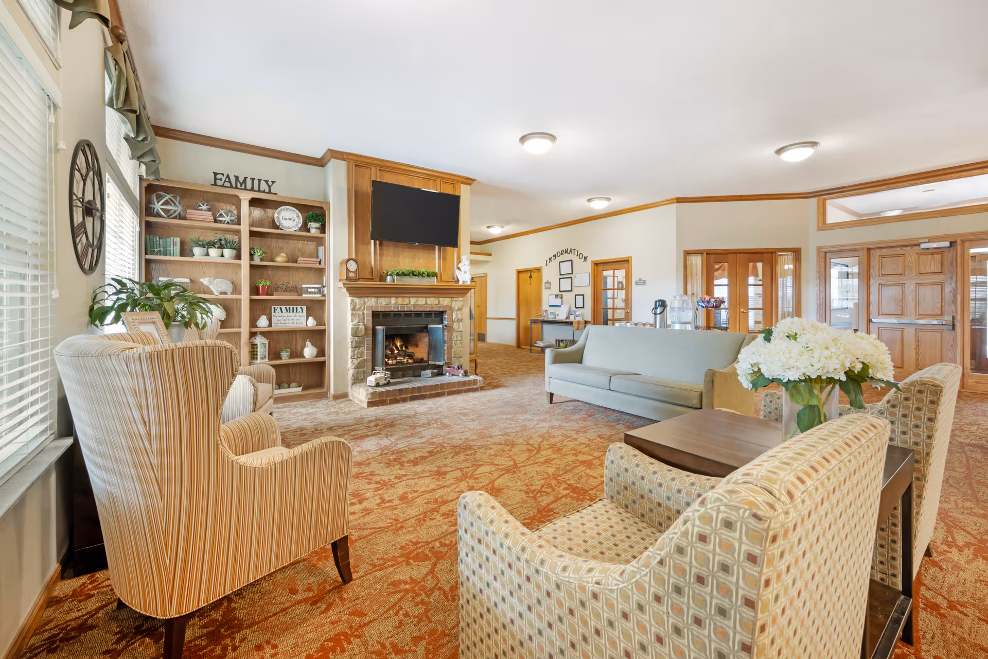 A cozy living room area in a senior living facility with patterned armchairs, a light blue sofa, a wooden coffee table with a vase of white flowers, a brick fireplace with a TV mounted above it, and a wooden bookshelf decorated with plants and family-themed decor. The room has large windows with blinds, warm lighting, and carpeted floors with a red and beige pattern.