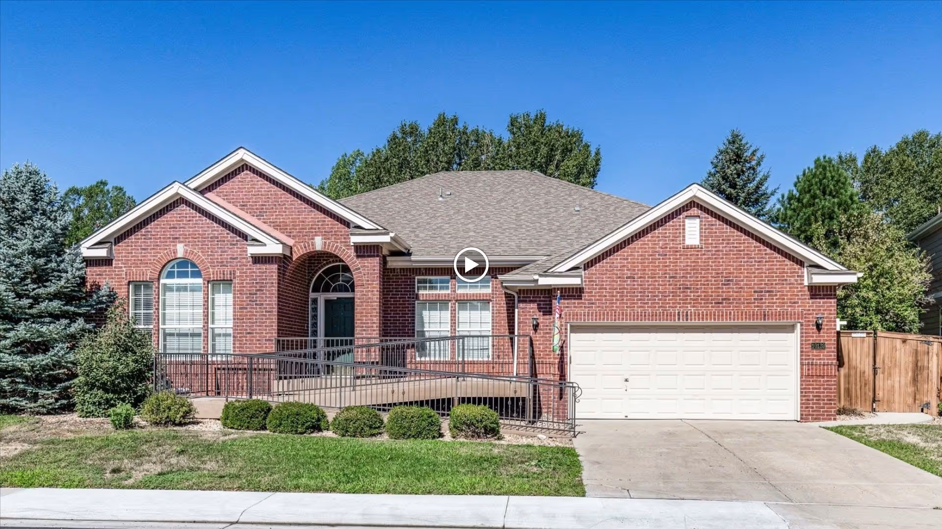Front exterior view of a single-story brick house with a gray shingled roof, a wheelchair accessible ramp leading to the front door, a two-car garage, and well-maintained landscaping including bushes and trees under a clear blue sky.