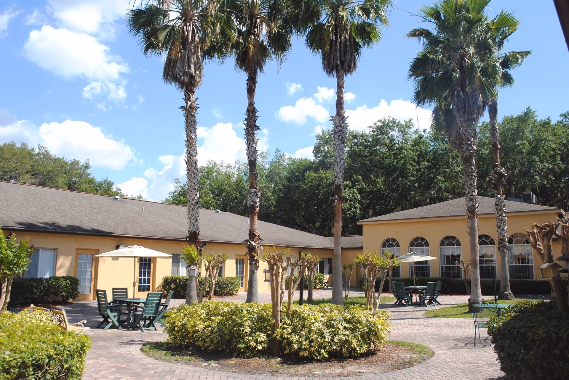 Outdoor courtyard area at The Club at St Cloud featuring tall palm trees, green shrubs, paved walkways, and patio tables with umbrellas and chairs under a bright blue sky with some clouds.