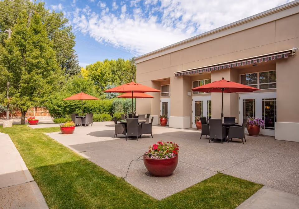 Outdoor patio area with several round tables and black wicker chairs, each table shaded by a red umbrella. Large red flower pots with colorful flowers are placed around the patio. The patio is adjacent to a beige building with large windows and doors, and there is green grass and trees surrounding the area under a partly cloudy sky.