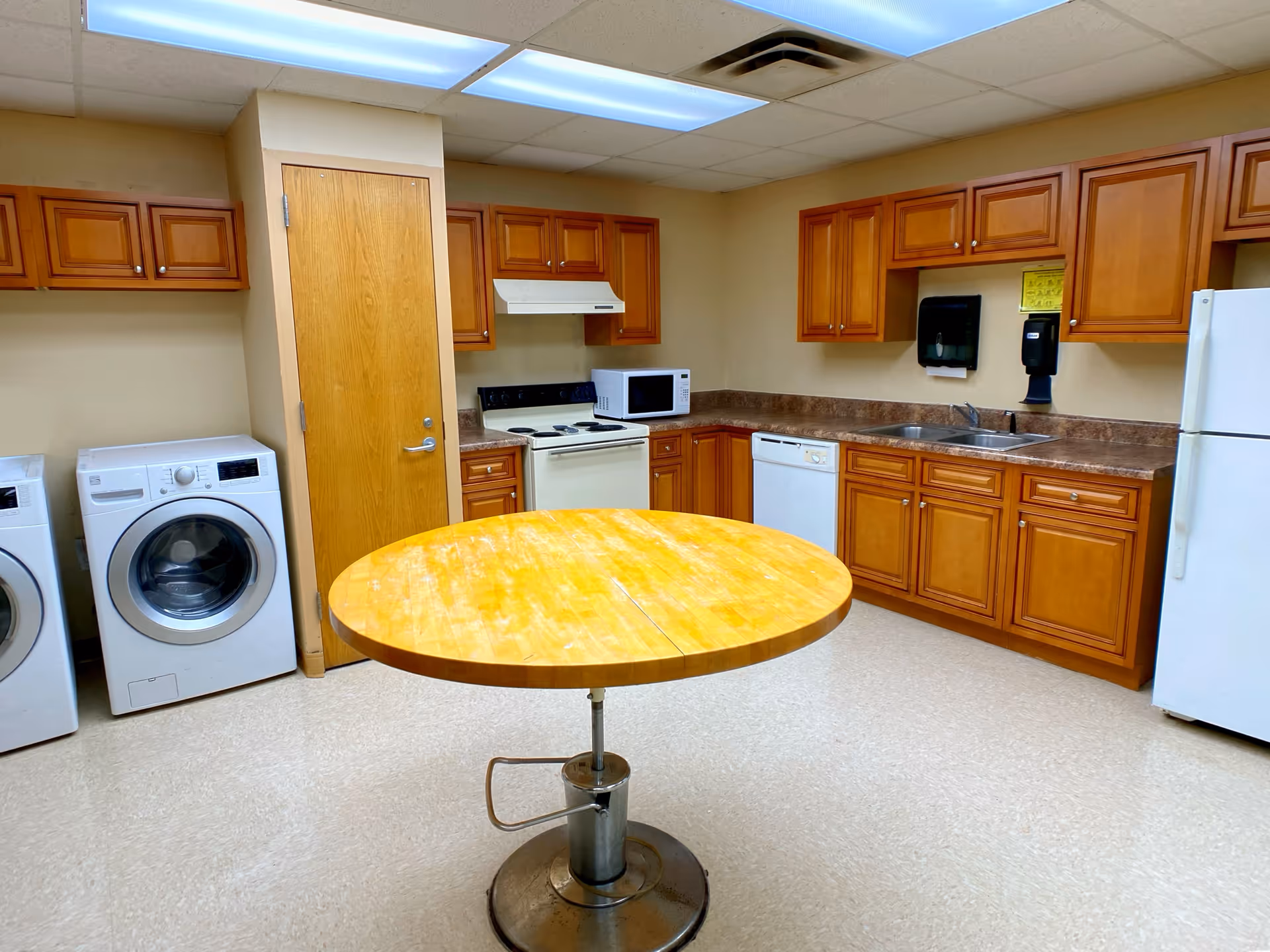 A kitchen area with wooden cabinets, a white refrigerator, dishwasher, microwave, stove, and a round wooden table in the center. To the left, there is a washer and dryer set next to a wooden door. The room has a tiled floor and fluorescent ceiling lights.