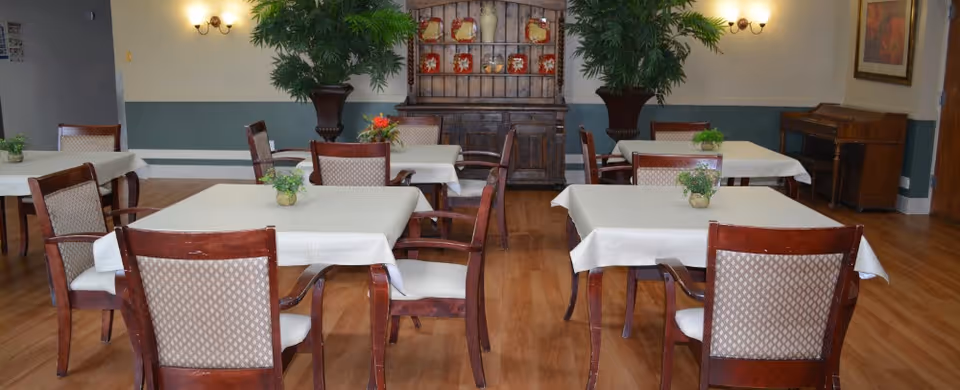 A dining room with several tables covered in white tablecloths, each surrounded by wooden chairs with cushioned seats and backs. Small potted plants decorate the tables. The room has wooden flooring, two large potted plants near the back wall, a wooden cabinet with decorative plates, wall sconces providing light, and a piano in the corner.