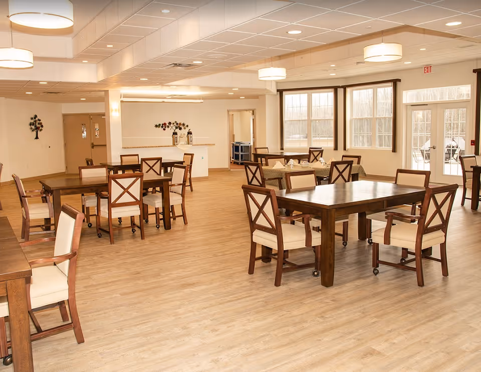 A spacious dining room in The Lakes at Jefferson facility with multiple wooden tables and cushioned chairs arranged neatly. The room has large windows allowing natural light to fill the space, light wood flooring, and modern ceiling lights. There is a counter area in the back with decorative wall art and a door leading to another room.