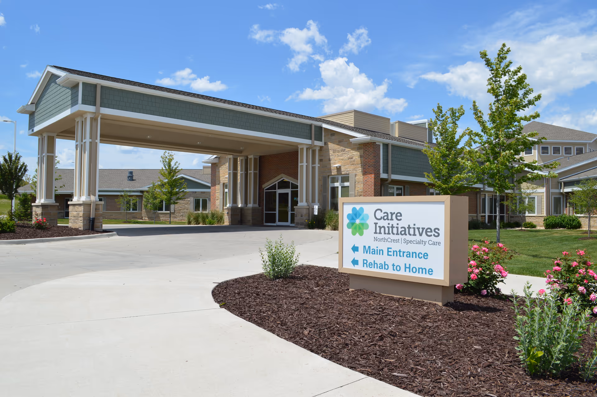 Exterior front entrance of a senior care facility with a covered porte-cochere, landscaped grounds, and a Care Initiatives sign pointing to the main entrance and rehab to home.