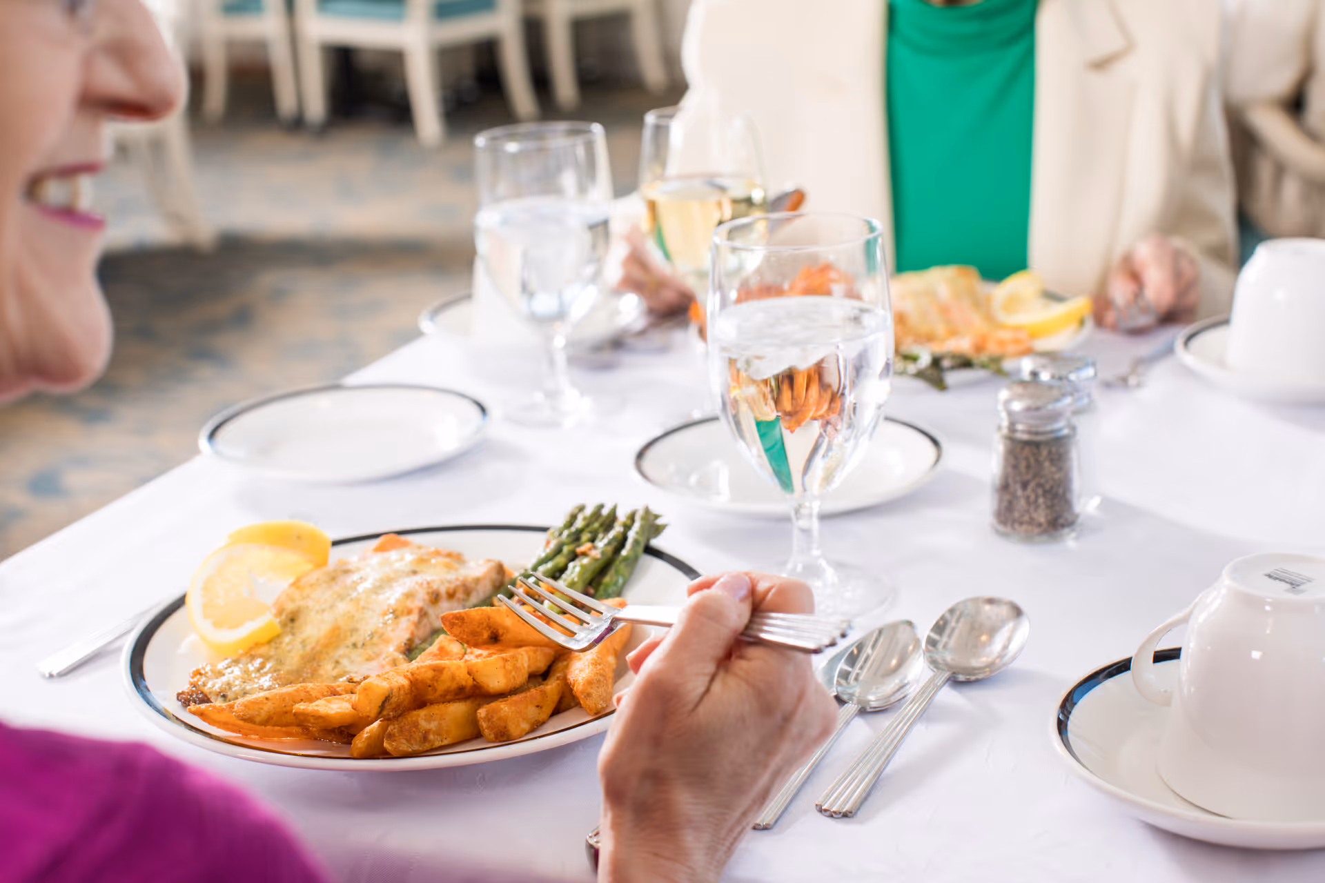 Two people seated at a dining table with white tablecloths, enjoying a meal that includes fish, potato wedges, and asparagus. There are glasses of water and wine, silverware, and cups on the table.