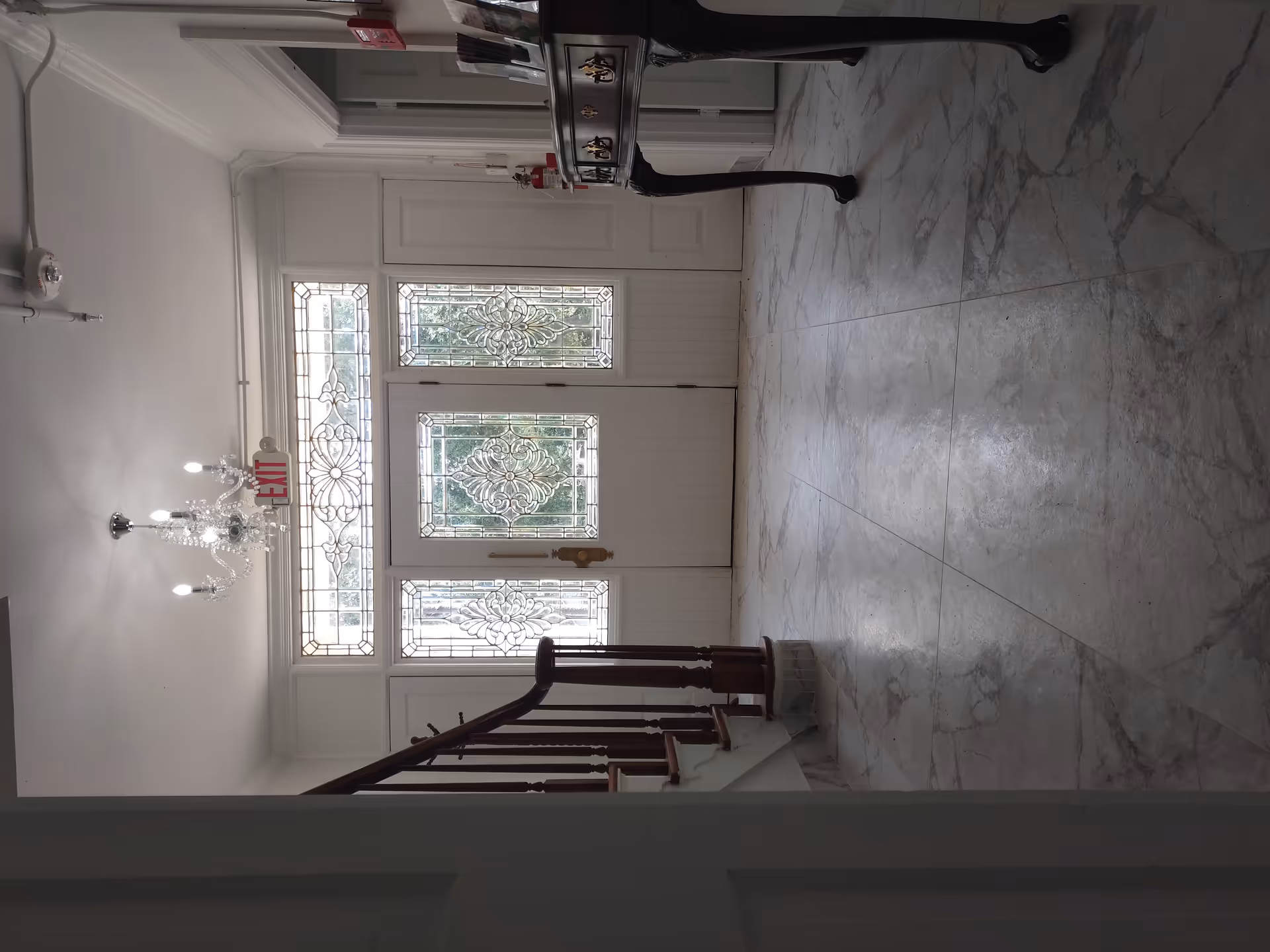 Interior view of an entryway with a decorative glass front door and sidelights, a chandelier hanging from the ceiling, a wooden staircase with a handrail on the left, and a dark wooden console table on the right with brochures on top.