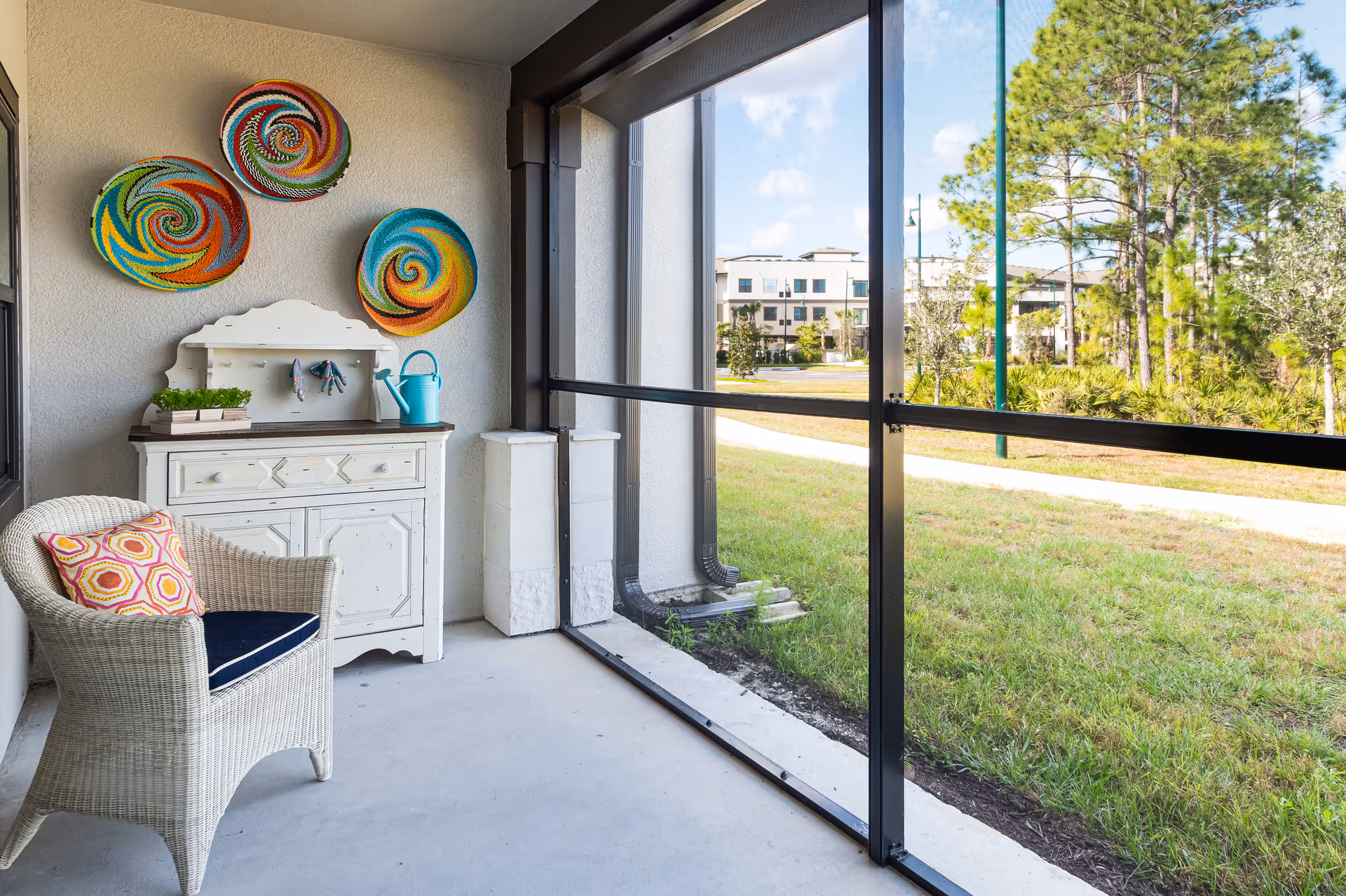 A screened-in porch area with a white wicker chair featuring a colorful cushion, a white wooden cabinet with gardening tools and a watering can on top, and three colorful circular wall decorations. Outside the screened porch is a grassy area with trees and a building in the background under a partly cloudy sky.